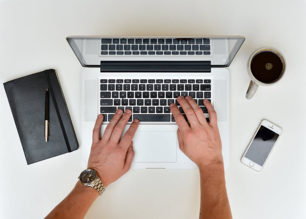 Hands typing on a laptop beside a notebook, a phone and a cup of coffee